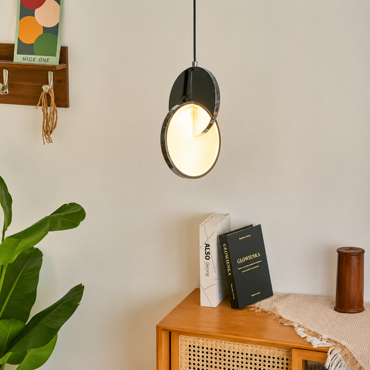 A modern pendant light with a matte black circular shade and a warm glowing white inner ring, hanging in a styled room above a wooden side table with books and a plant.