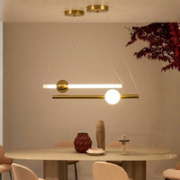 Modern linear pendant light with a horizontal bar in brushed brass and a glowing white spherical bulb, hanging over a dining table in a minimalist interior.