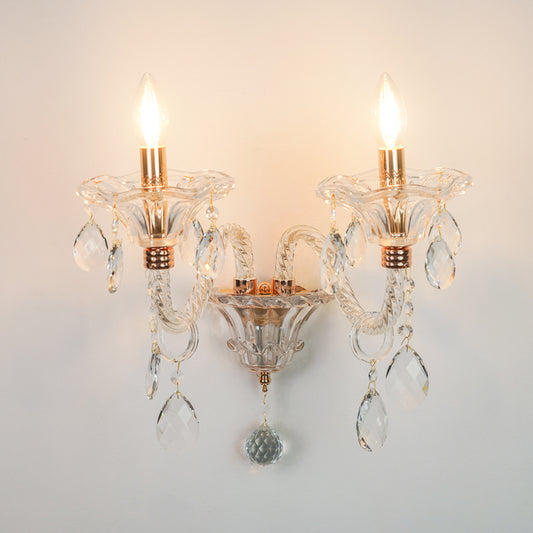 A two-light wall sconce with clear glass crystal arms and hanging teardrop prisms, featuring warm brass/gold-toned accents and illuminated bulbs, mounted against a plain light wall.