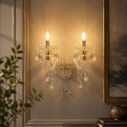 A two-light crystal wall sconce with gold-toned metal arms and hanging clear teardrop prisms, mounted on a beige wall in an interior setting with a framed print and plant partially visible.