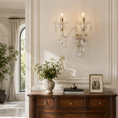 A double-tier crystal wall sconce with gold-toned metal arms and hanging clear glass teardrop prisms, mounted on a cream paneled wall above a dark wood console table in a styled interior.