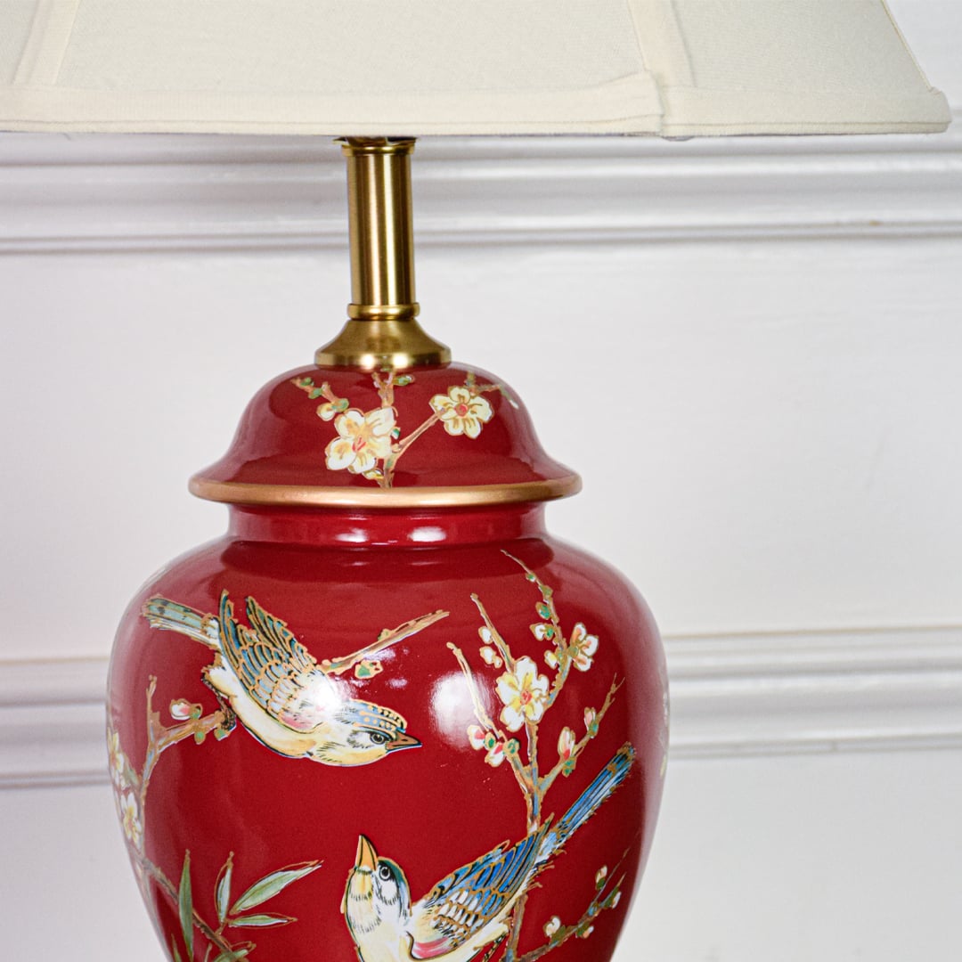 Closeup of red ceramic table lamp highlighting hand painted bird floral detailing smooth glazed finish and gold metal base.