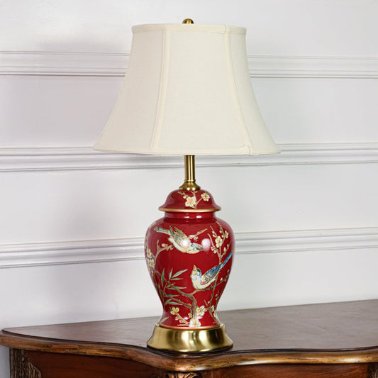 Decorative red ceramic table lamp with artistic bird floral design gold metal base and white fabric shade on off white background.