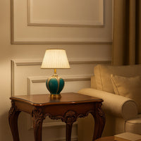 Turquoise ceramic table lamp with pleated white shade glowing on wooden side table for elegant bedroom interior lighting.