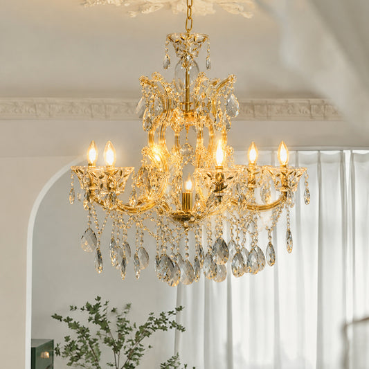 A gold chandelier with clear crystal hanging drops and candle-style lights, shown hanging in an elegant interior room with white curtains and a decorative archway.