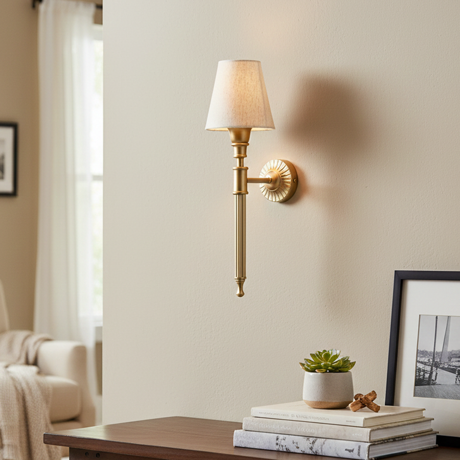 Classic gold metal torch wall sconce lamp with a cream fabric cone shade casting a warm light over a home office desk.