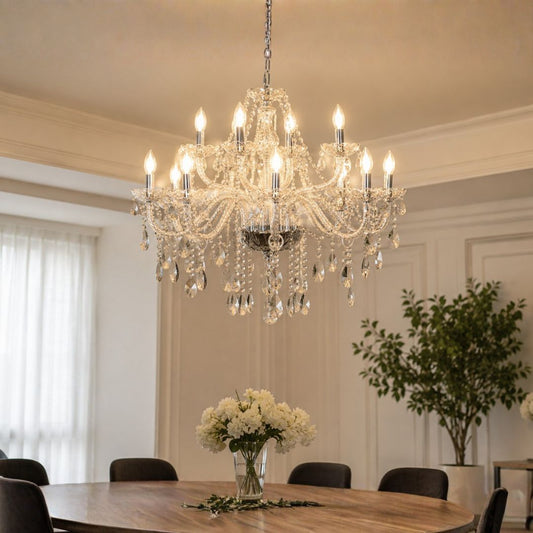 A large multi-tier crystal chandelier with lit candle-style bulbs hanging over a dining table in a bright, elegant room with white walls and a potted plant in the background.