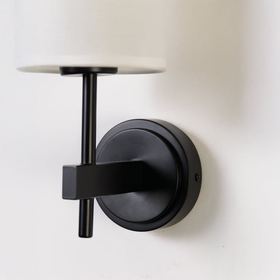 Closeup of black metal wall light with tall white fabric shade showing premium detailing for modern indoor lighting in stylish home interiors.