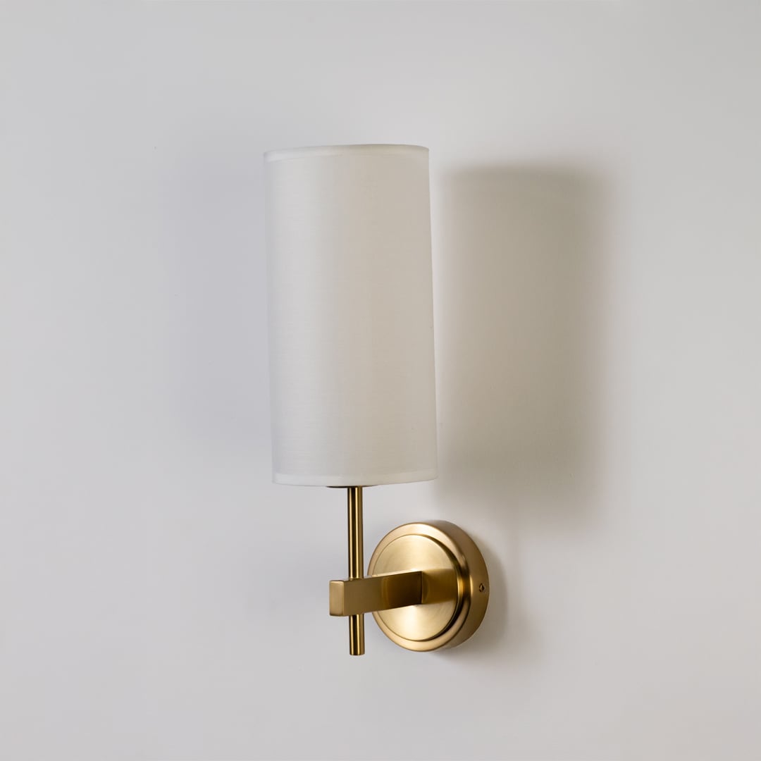 Gold metal wall light with tall white fabric shade shown on a clean white background with light off highlighting its elegant modern shape.