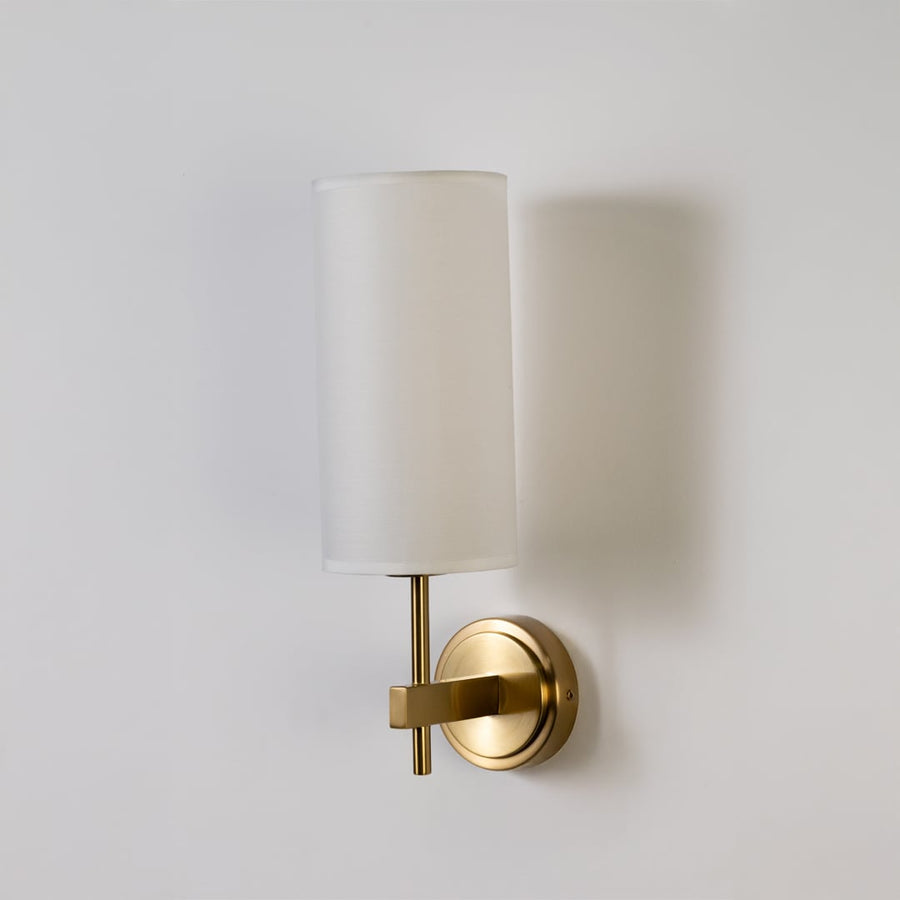 Gold metal wall light with tall white fabric shade shown on a clean white background with light off highlighting its elegant modern shape.