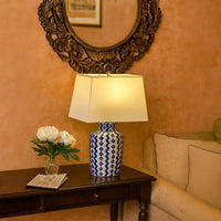 Blue patterned lamp with yellow shade on side table with light on and tan wall with carved mirror background for vibrant décor.