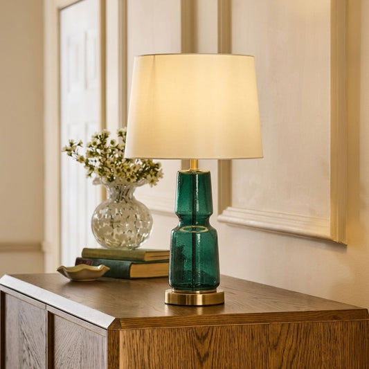 A table lamp with a textured teal-green glass base and a warm cream-colored fabric drum shade, illuminated and placed on a wooden console table in a home interior setting.