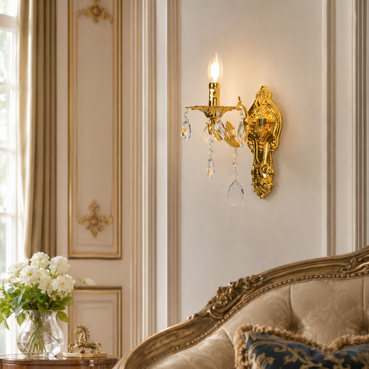 An ornate gold wall sconce with clear crystal droplets, mounted on a cream paneled wall in a luxurious room setting with a tufted sofa and flowers.