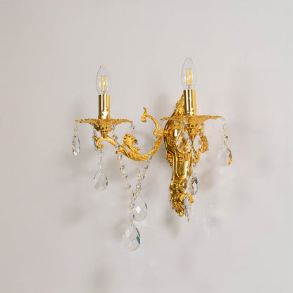 Golden wall light with crystal drops elegant twin candle style wall mounted decorative fixture on off white background.