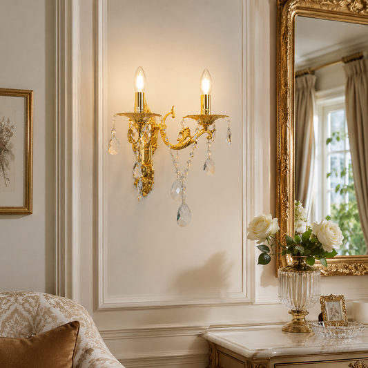An ornate gold wall sconce with hanging clear crystal drops, mounted on a cream paneled wall in a luxurious room setting with a gold mirror and a marble console table.