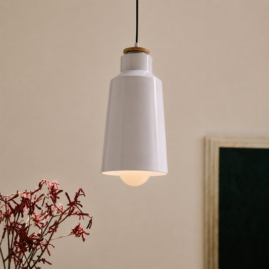 A modern white pendant light with a tapered bell-shaped shade, a light wood accent ring near the top, and a black hanging cord, shown against a warm beige wall with a blurred framed picture and dried red flowers in the foreground.