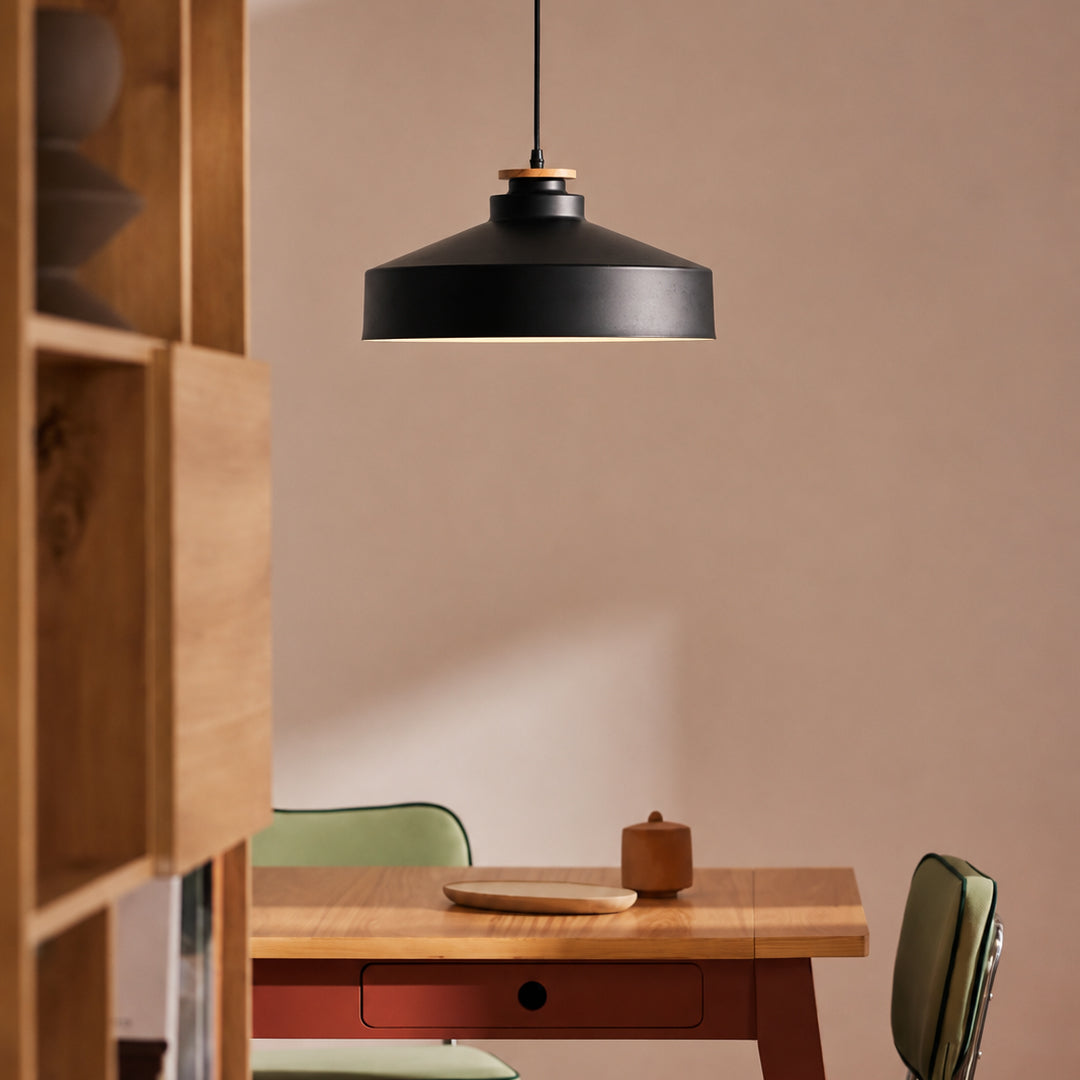 A matte black pendant light with a warm wood accent ring hangs over a wooden dining table in a minimalist room with beige walls.