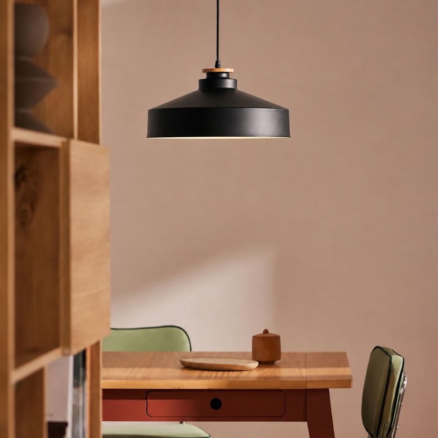 A matte black pendant light with a warm wood accent ring hangs over a wooden dining table in a minimalist room with beige walls.