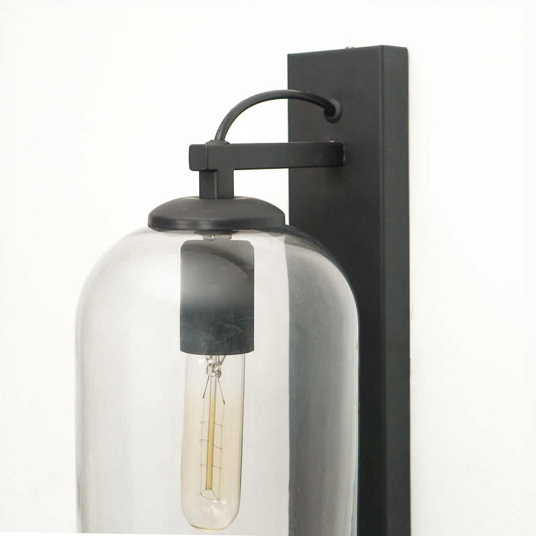 A modern wall-mounted light fixture with a matte black rectangular backplate, curved arm, and clear glass dome shade housing an exposed filament bulb, shown against a plain white background.