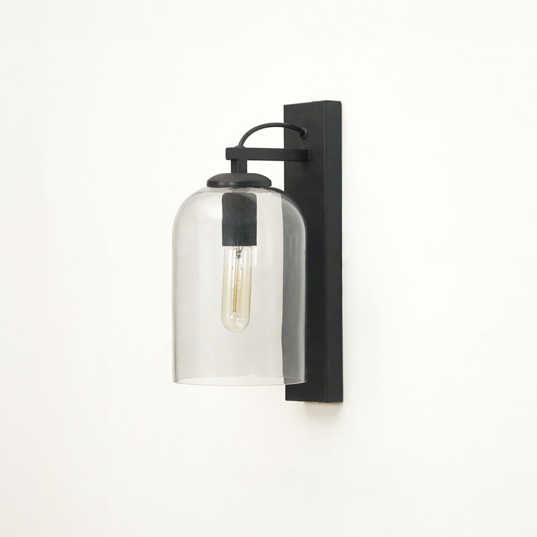 A modern wall sconce light fixture with a matte black rectangular wall mount, curved top bar, and a clear cylindrical glass shade enclosing an exposed Edison-style bulb, shown against a plain white wall.