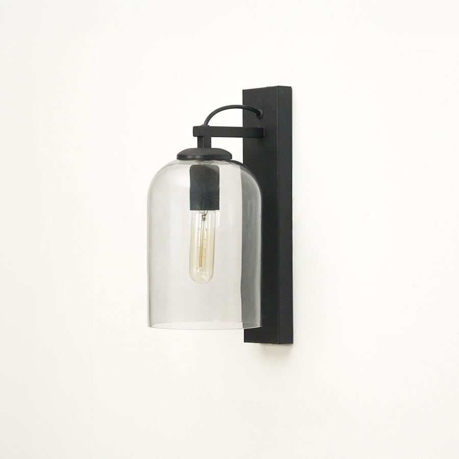 A modern wall sconce light fixture with a matte black rectangular wall mount, curved top bar, and a clear cylindrical glass shade enclosing an exposed Edison-style bulb, shown against a plain white wall.