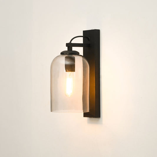A modern industrial wall sconce with a black metal bracket and a clear cylindrical glass shade housing a glowing warm filament bulb, mounted on a plain off-white wall.