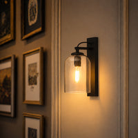 A wall-mounted industrial-style light fixture with a black metal bracket and a clear glass bell shade, glowing with a warm light on a beige wall next to framed artwork.