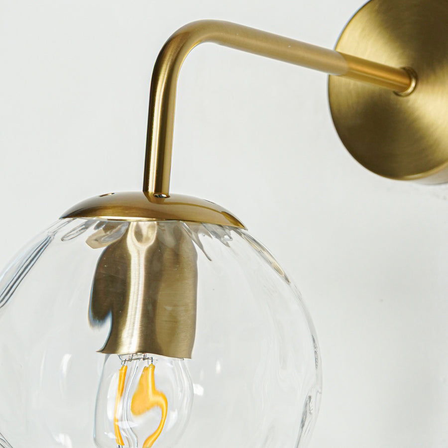 Modern brass wall light with clear textured glass globe and elegant curved arm for luxury interiors.