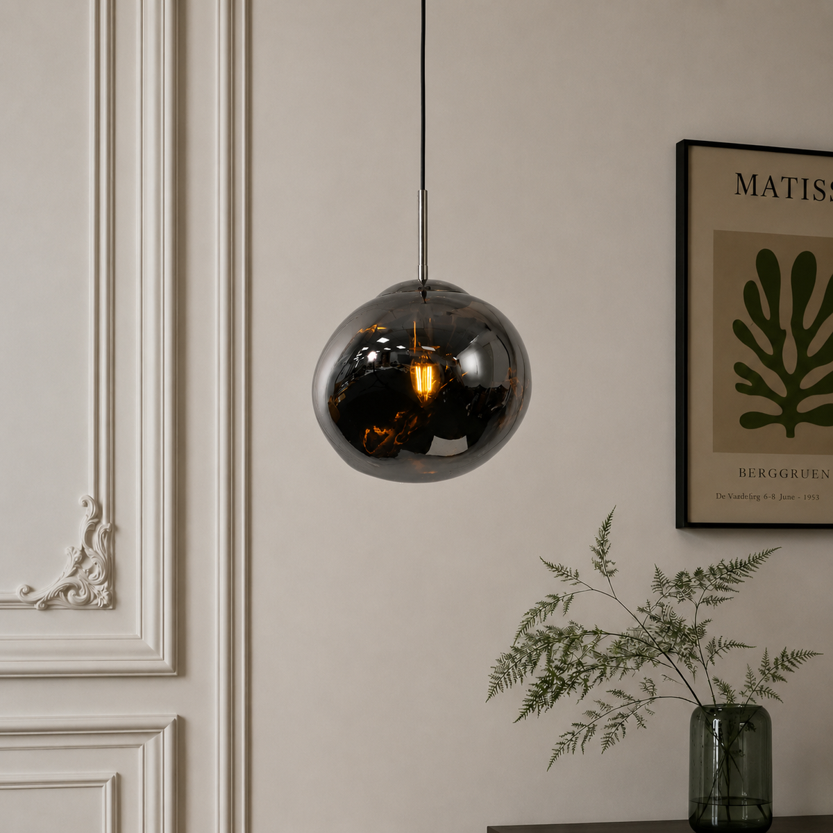 A spherical smoked glass pendant light hangs from a black cord against a beige wall in a styled interior setting with a framed print and a green glass vase.