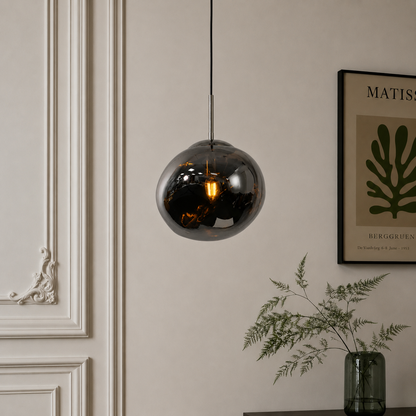A spherical smoked glass pendant light hangs from a black cord against a beige wall in a styled interior setting with a framed print and a green glass vase.