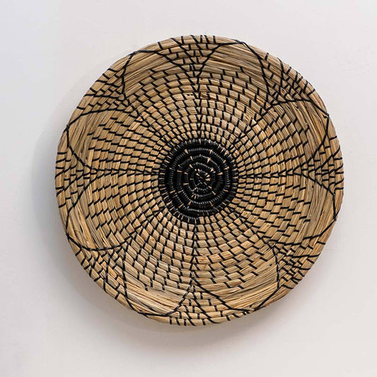Round handwoven seagrass wall basket with black flower motif for bohemian living room and bedroom accent on white background.
