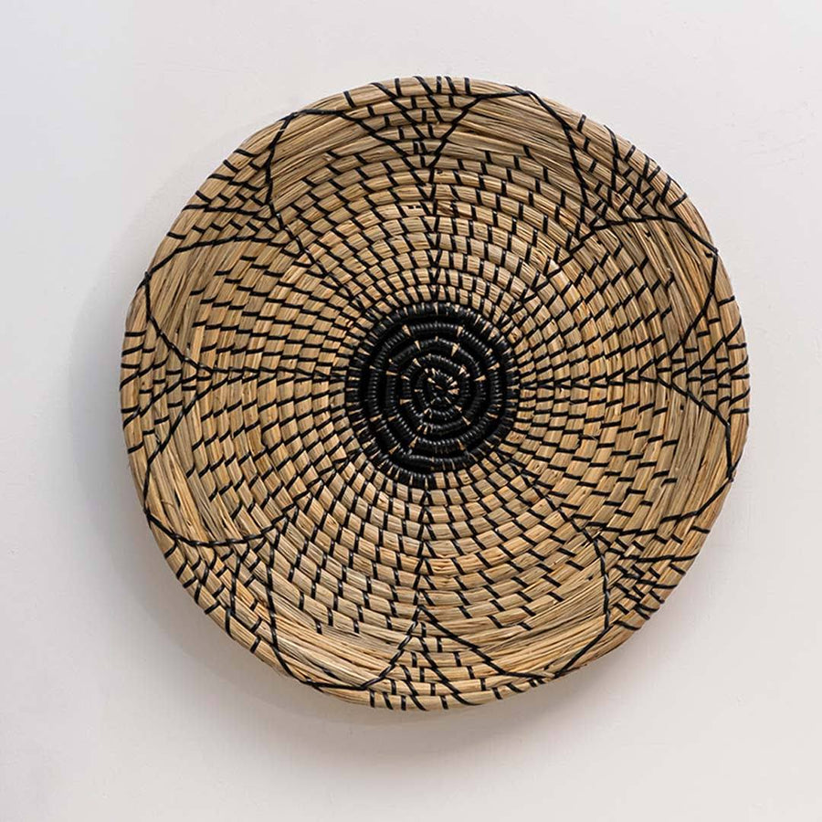 Round handwoven seagrass wall basket with black flower motif for bohemian living room and bedroom accent on white background.