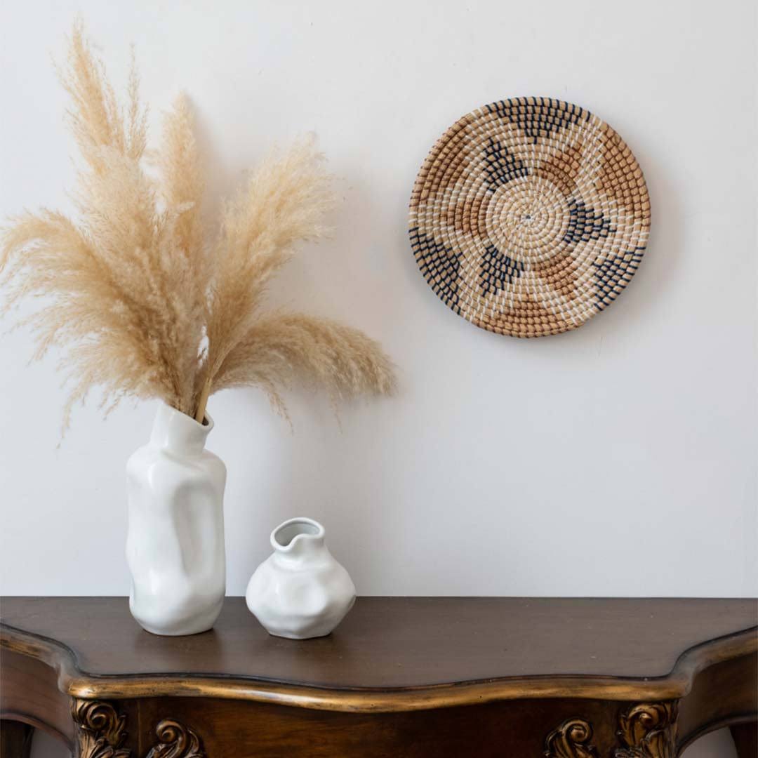 Geometric woven seagrass wall basket plate hanging above a wooden console table decorated with vases and pampas grass.