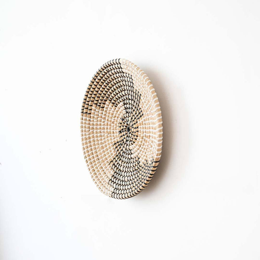 Side angle view of a spiral pattern black woven seagrass wall hanging basket plate showing depth on a white wall.