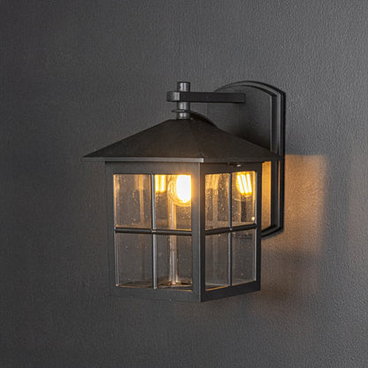 Modern black metal outdoor wall lantern turned on against dark background showcasing warm glow through clear glass panels.