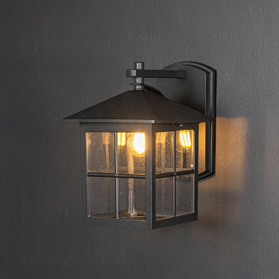 Modern black metal outdoor wall lantern turned on against dark background showcasing warm glow through clear glass panels.