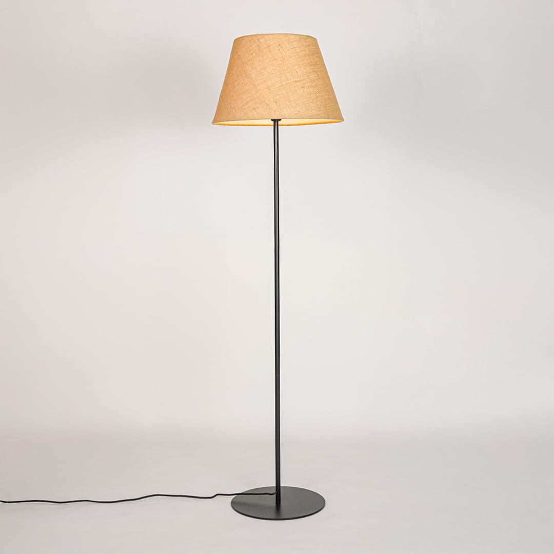 Black standing floor lamp illuminated through beige fabric shade casting soft warm glow on white backdrop.