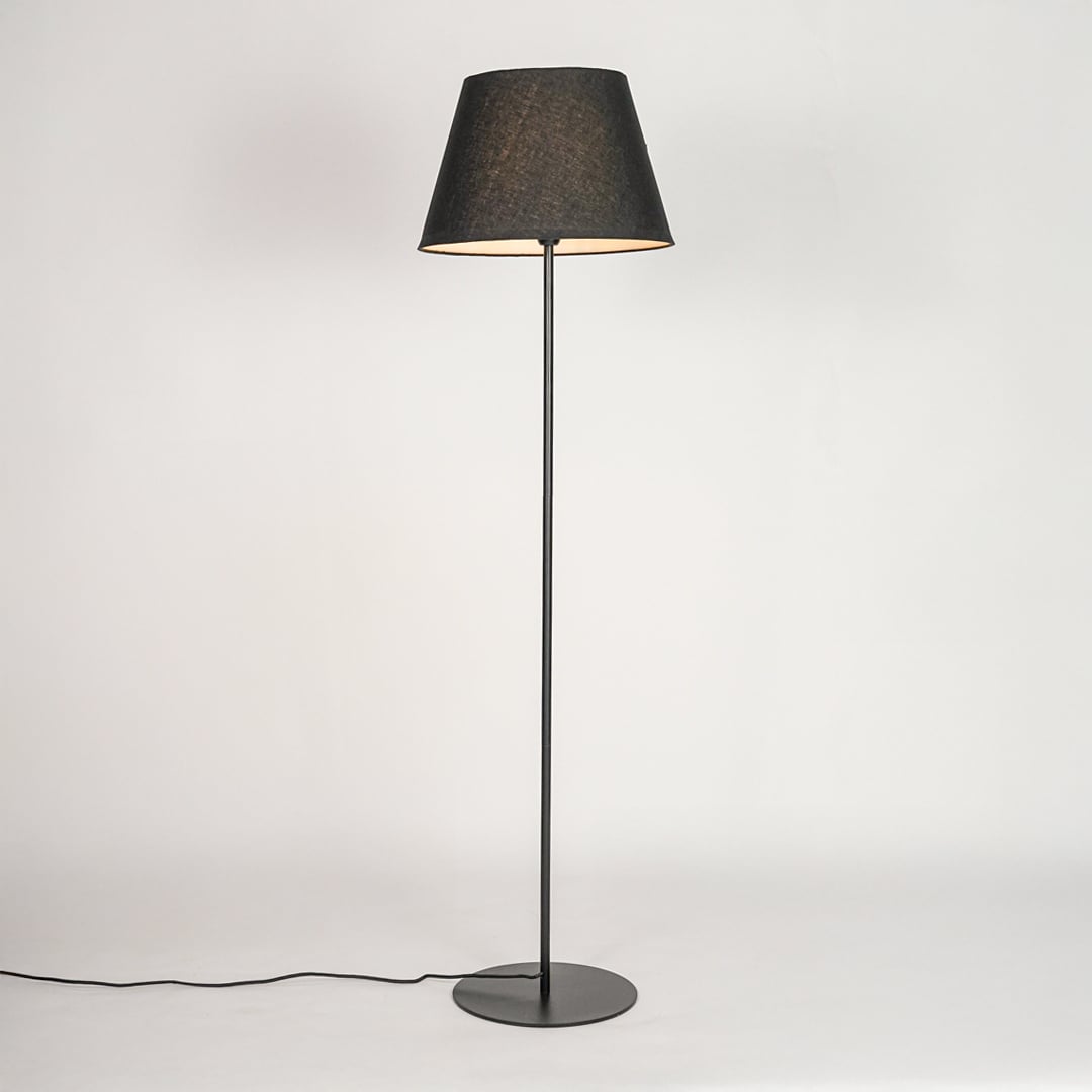 Black floor lamp illuminated through dark grey shade casting soft warm glow against white backdrop.