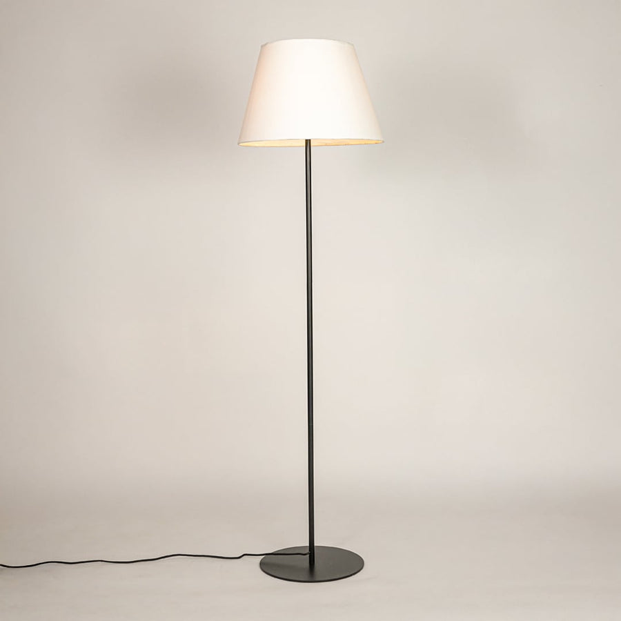 Black metal floor lamp glowing warmly through white fabric shade on white background for cozy home Decor.
