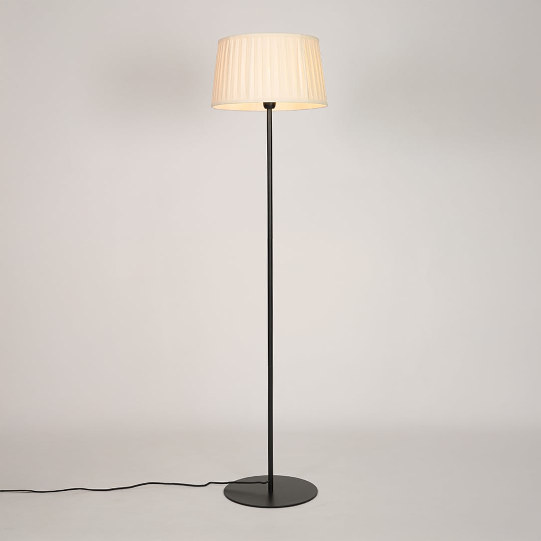 Black metal floor lamp glowing warmly through pleated cream fabric shade on white background for cozy home Decor.