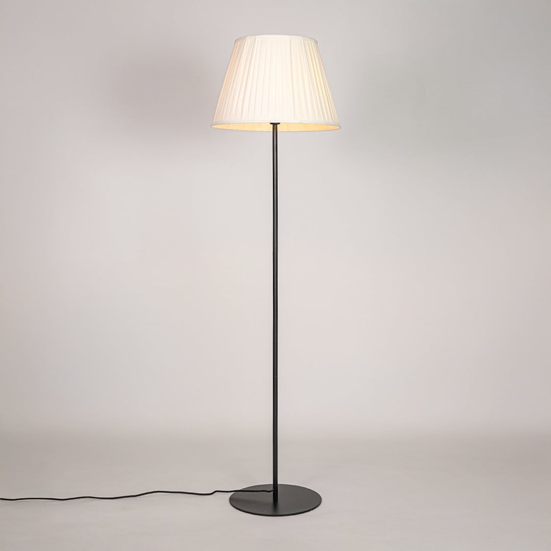 Black floor lamp illuminated with warm glow through pleated cream shade against bright white studio backdrop.
