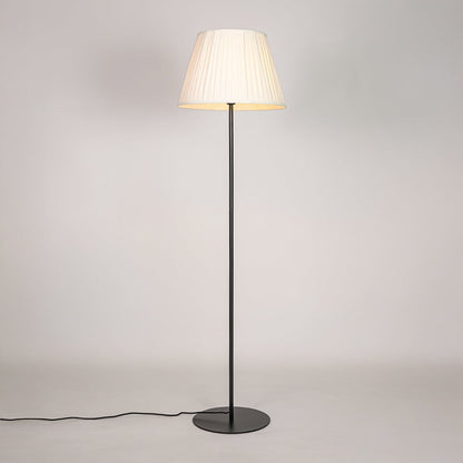Black floor lamp illuminated with warm glow through pleated cream shade against bright white studio backdrop.