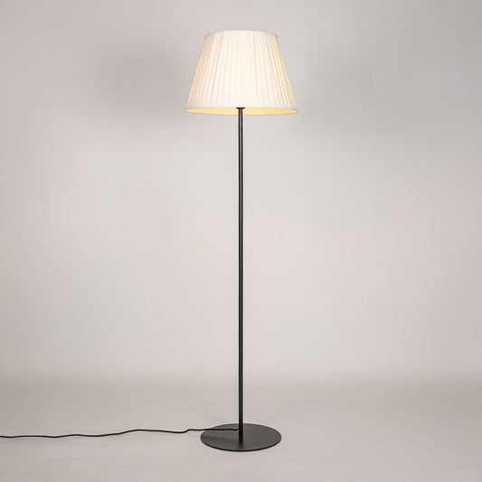 Black floor lamp illuminated with warm glow through pleated cream shade against bright white studio backdrop.