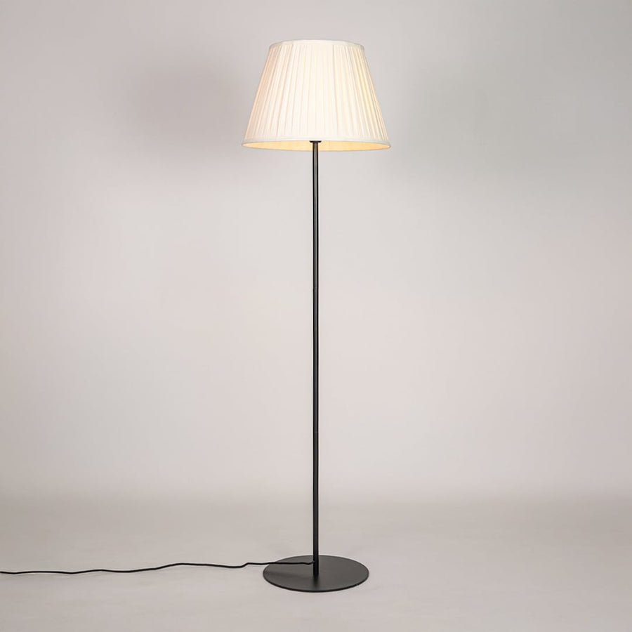 Black floor lamp illuminated with warm glow through pleated cream shade against bright white studio backdrop.