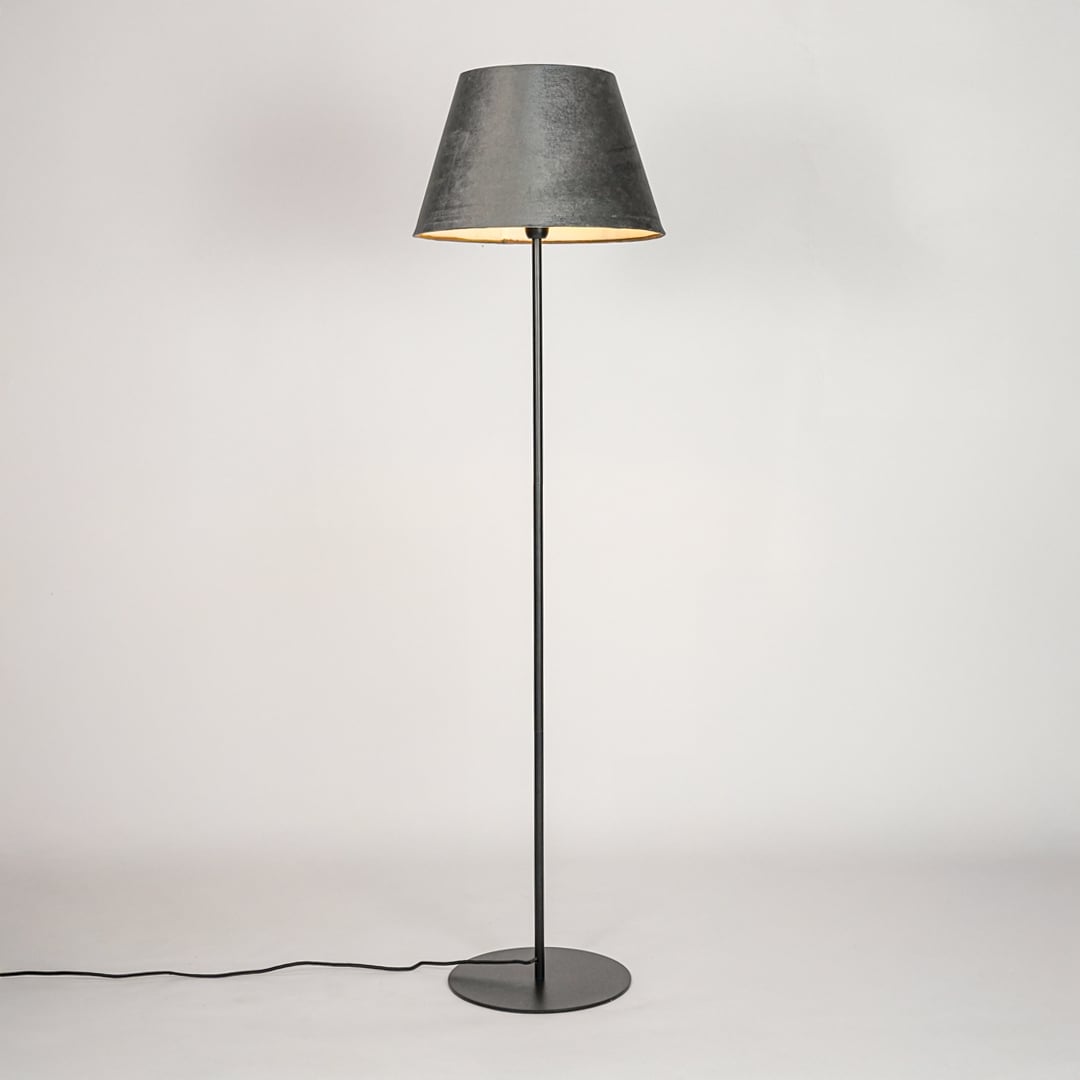 Black floor lamp illuminated through grey velvet shade casting warm cozy glow against white backdrop.