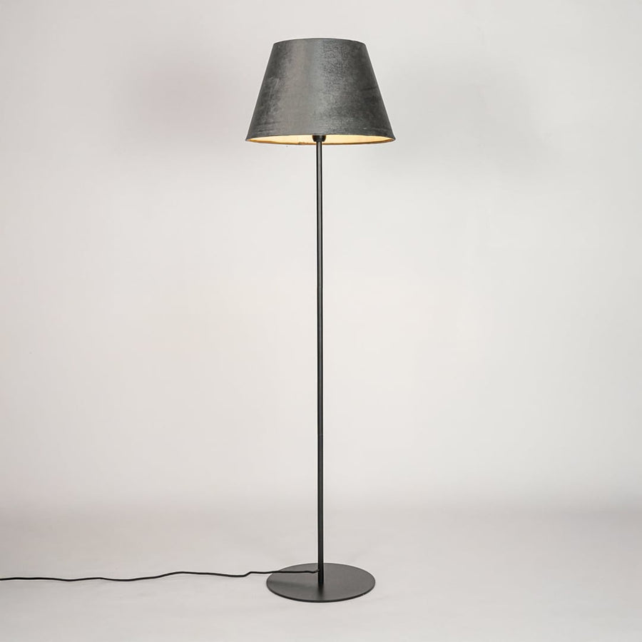 Black floor lamp illuminated through grey velvet shade casting warm cozy glow against white backdrop.