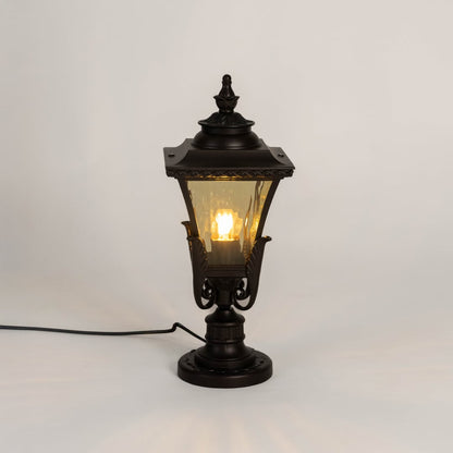 Designer outdoor black metal pillar lamp glowing with warm amber glass shade perfect for patio terrace and driveway.