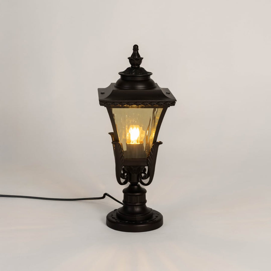 Designer outdoor black metal pillar lamp glowing with warm amber glass shade perfect for patio terrace and driveway.