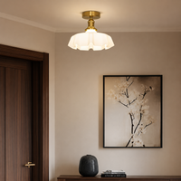 A modern semi-flush ceiling light with a white textured glass shade and a gold brass fixture, mounted on a beige wall in a styled interior next to a dark wood door and a framed floral print.