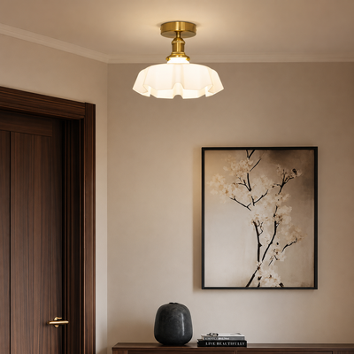 A modern semi-flush ceiling light with a white textured glass shade and a gold brass fixture, mounted on a beige wall in a styled interior next to a dark wood door and a framed floral print.